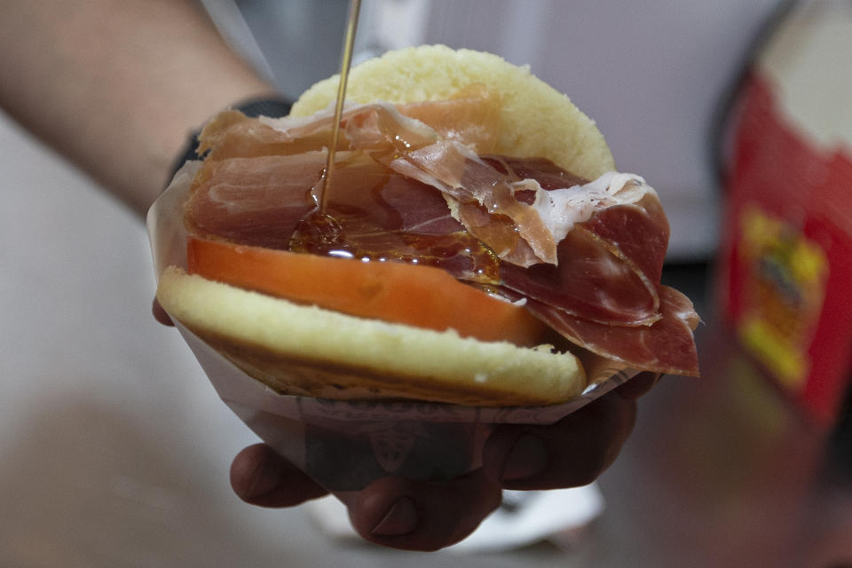 In this June 19, 2019 photo, chef and owner of the Arepa Ole restaurant, Edgar Rodriguez from Venezuela, prepares an Iberia Arepa with serrano ham, tomato and olive oil, in Madrid, Spain. The story of the arepa begins before the arrival of Spanish colonizers. Indigenous chefs in Colombia and Venezuela ground white corn into round patties and baked them on clay griddles. (AP Photo/Paul White)