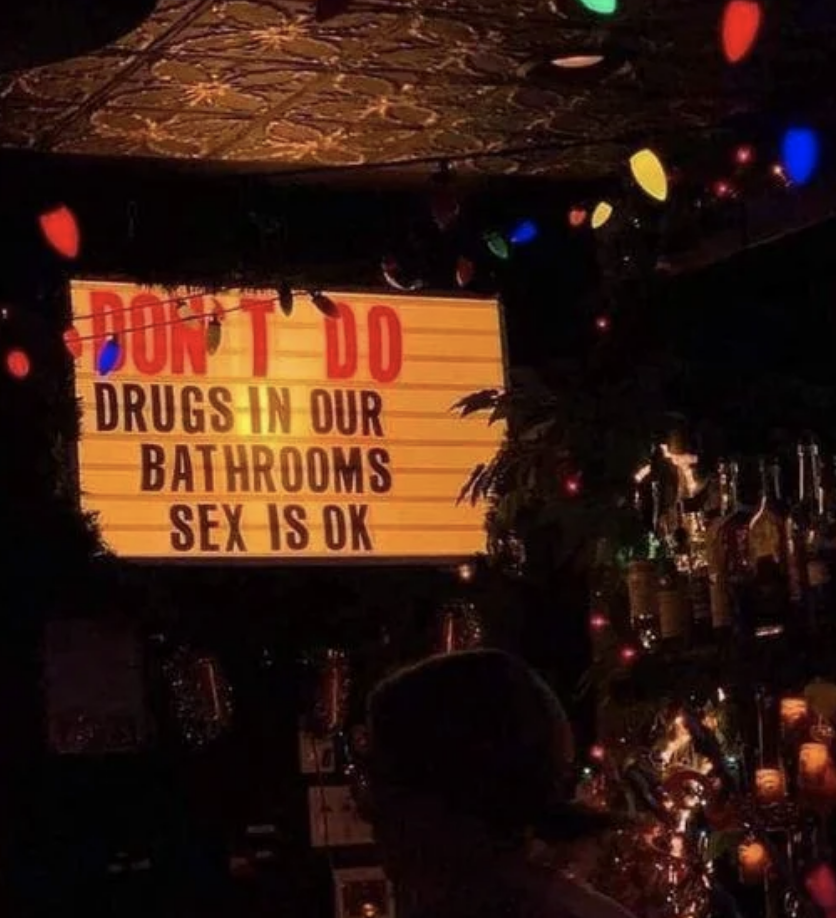 Sign in a bar reads "DON'T DO DRUGS IN OUR BATHROOMS, SEX IS OK" above a shelf of bottles