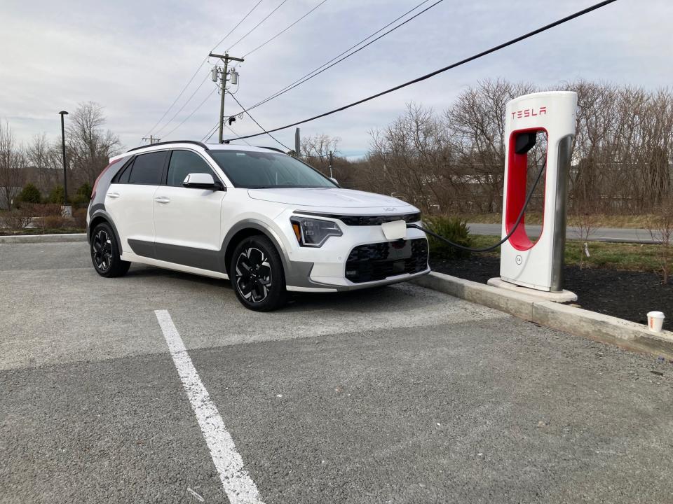 A white Kia Niro EV charges at a Tesla Supercharger in Brewster, NY, with clouds in the background.