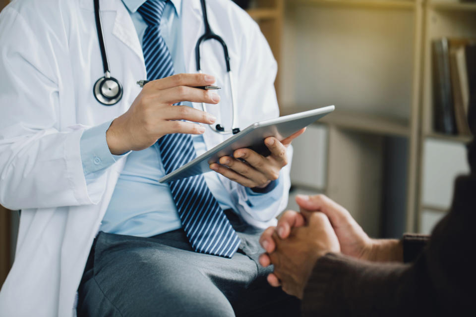 A doctor in a white coat holding a tablet consults with a patient who is clasping their hands together