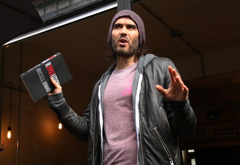 Russell Brand in a purple shirt and a grey hoodie and a beanie holding up both of his hands with a book in his right hand