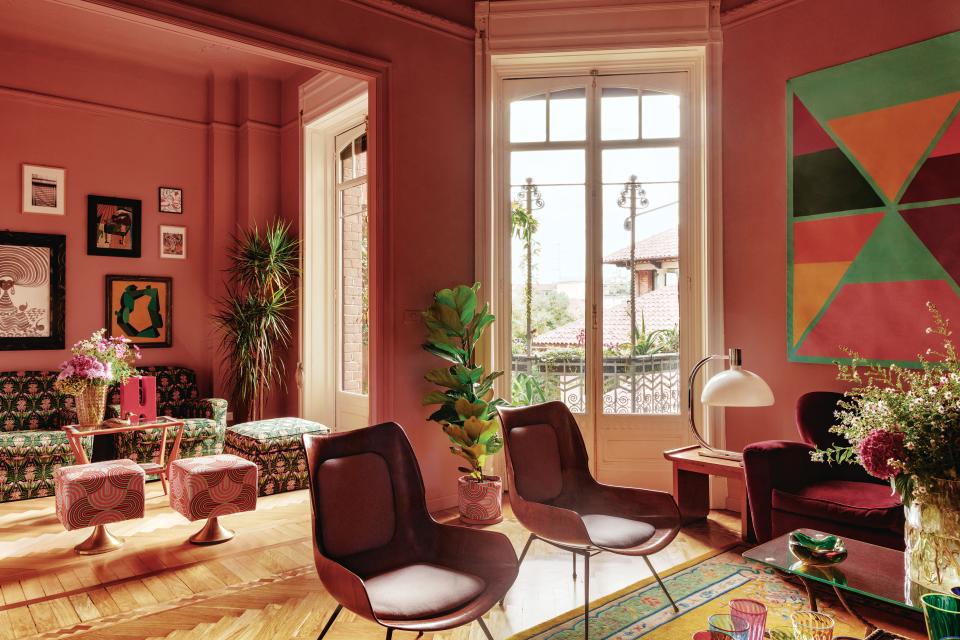 In the living room, a tapestry designed by Karim Noureldin hangs above a vintage 1950s sofa upholstered in red velvet. 
Carlo Ratti wooden chairs; vintage Franco Albini lamp on side table. In sitting area at back, the custom armchairs and ottoman wear a vintage print reissued by Prada, and the stools are covered in a La DoubleJ print.