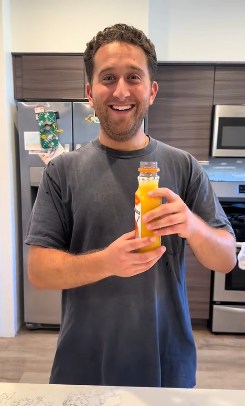 A man in a kitchen holds a bottle of orange juice, smiling at the camera. A refrigerator with magnets is visible in the background