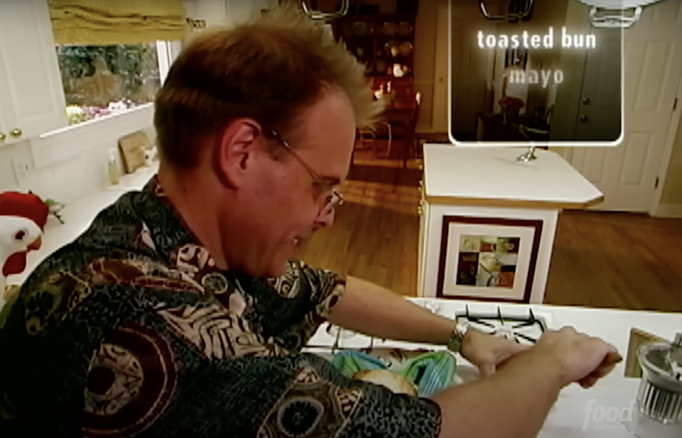 Man in patterned shirt toasting bun, condiments on counter, TV show scene from "Good Eats" with Alton Brown