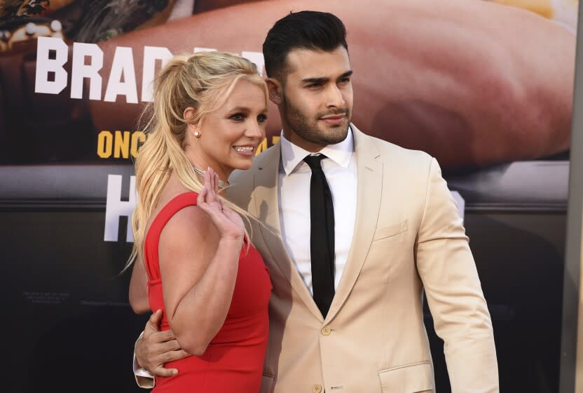 A man in a suit jacket holds his arm around a woman in a red dress as she smiles and waves