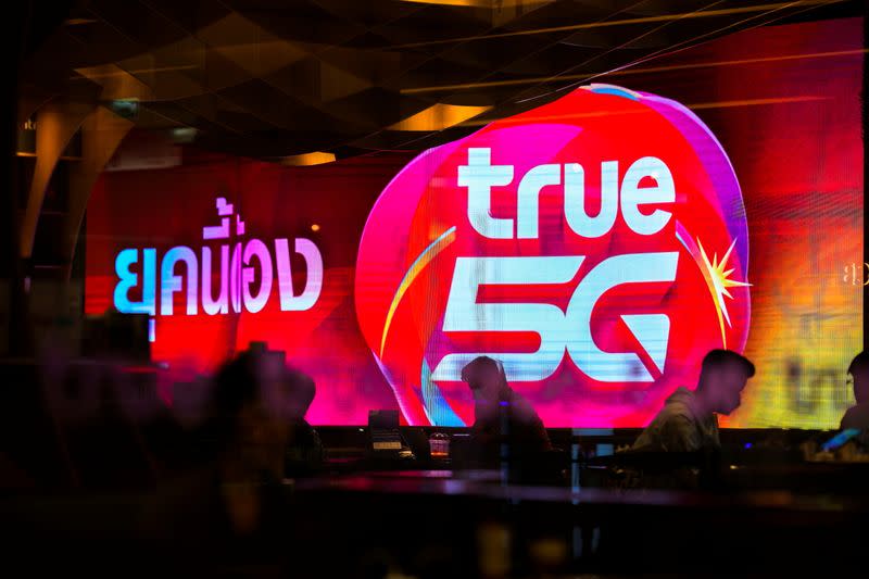 People are silhouetted in front of an electronic screen showing logo of the True Corp. at a department store in Bangkok