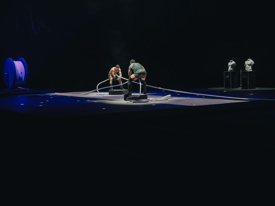 woo jin-yong and jung hae-min standing on two platforms in physical 100. they're each holding a thick rope attached on opposite sides to a spool, looking as if they're going to begin pulling on it