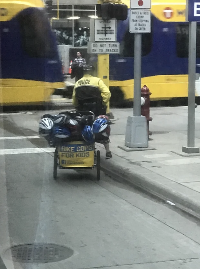 Person on bicycle with trailer labeled "Bike Cops for Kids" wearing a reflective jacket, passing a bus, near a "Do not turn on red" sign