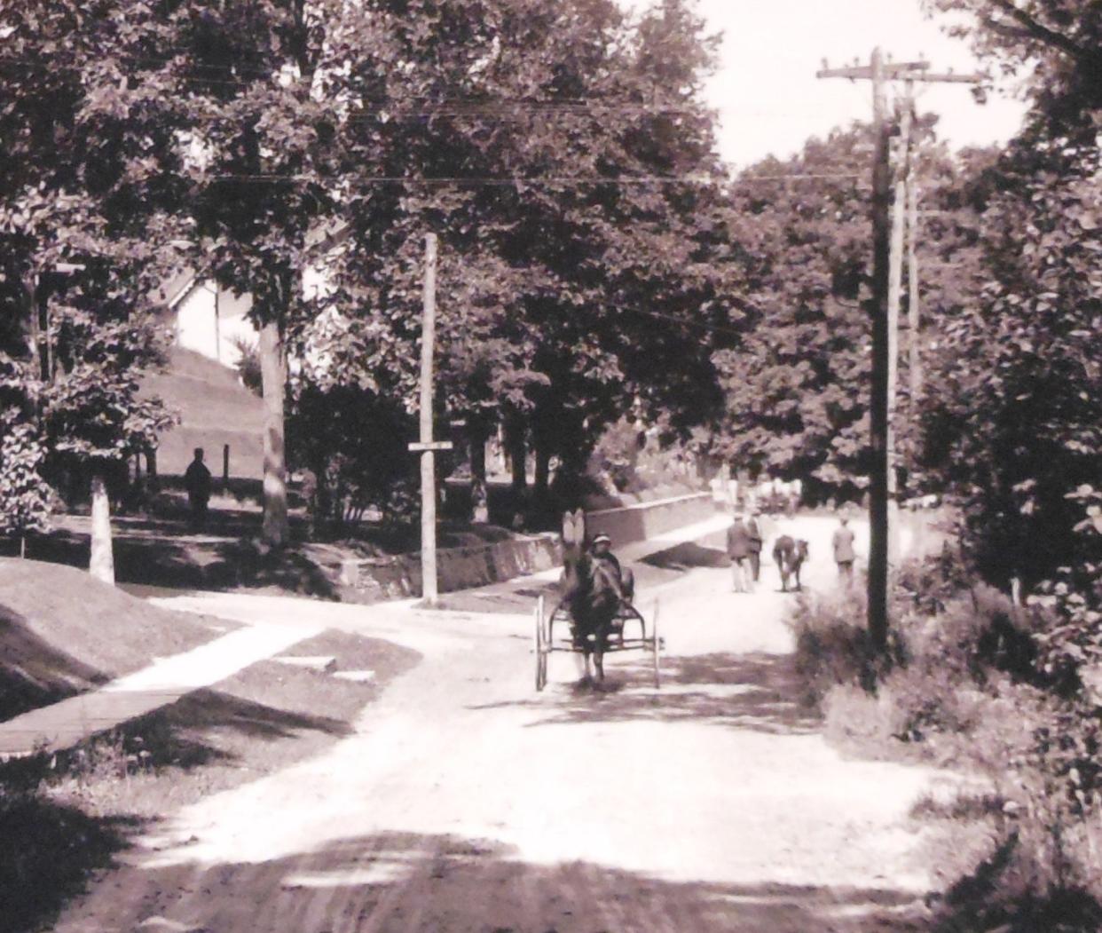 This is a scene on Bellemonte Avenue (later designated as part of Route 6) in Hawley, Pa., circa 1900-1910. The photo looks west down the hill one summer morning. At left in the shadow of the trees is a large man smoking a pipe, watching the world go by. This may be Charles Schumann, who lived on this corner with Woodland Avenue, at 254 Bellemonte Ave. Louis Hensel was the photographer.