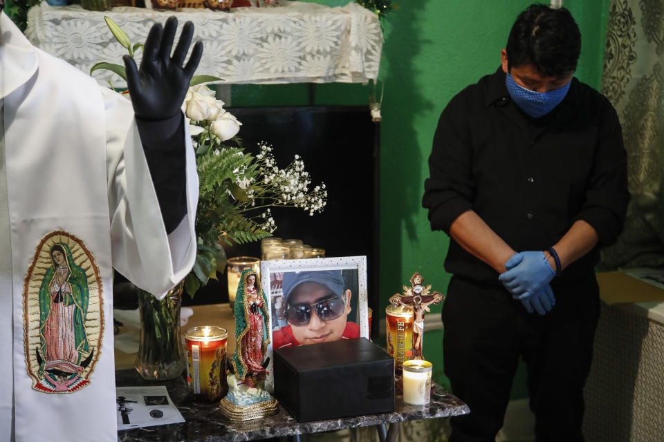 The Rev. Fabian Arias, left, performs an in-home service beside the remains of Raul Luis Lopez who died from COVID-19 the previous month as Lopez's cousin Miguel Hernandez Gomez, right, bows his head in prayer, Saturday, May 9, 2020, in the Corona neighborhood of the Queens borough of New York. (AP Photo/John Minchillo)