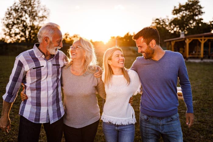 Older man, older woman, young woman, and young man stand smiling and embracing outdoors at sunset