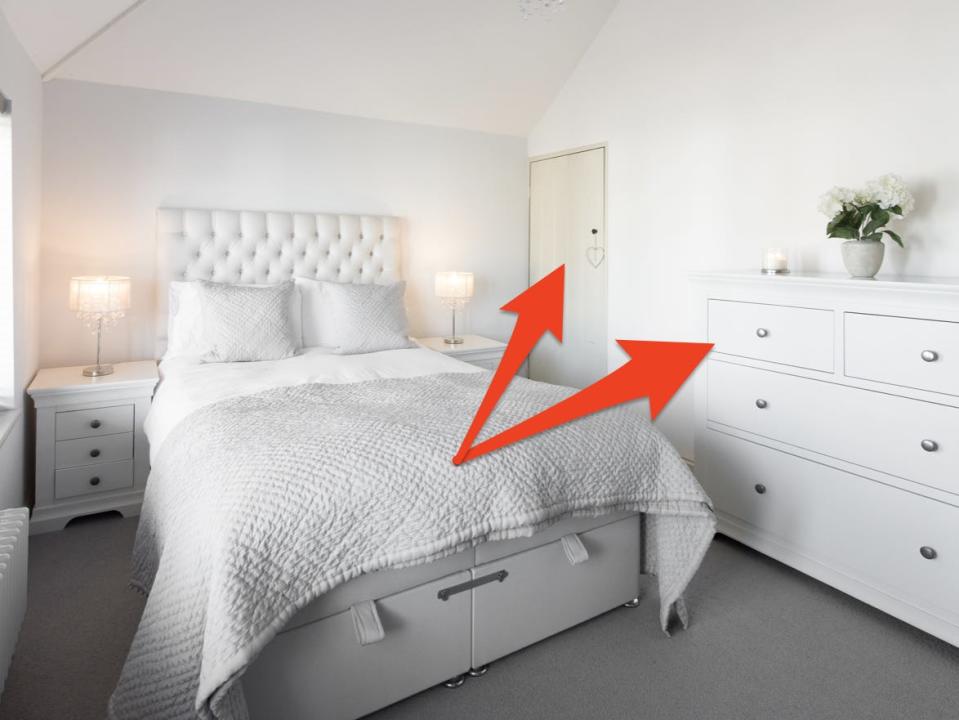 Bedroom with white sheets, white dresser, and white walls and door with red arrows pointing to dresser and door