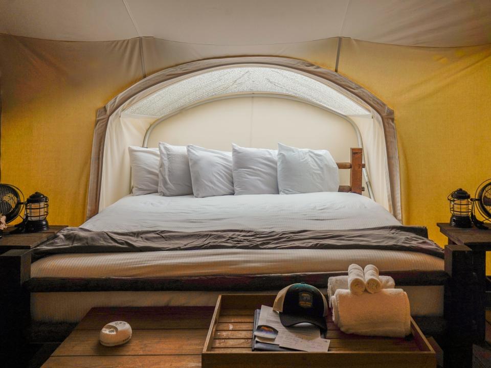 Inside a white tent with a white bed behind a wooden chest. There are night stands with lanterns on either side.