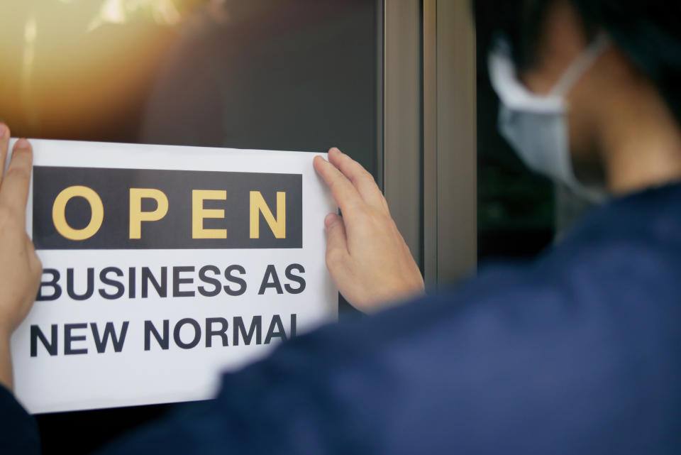 Reopening for business adapt to new normal in the novel Coronavirus COVID-19 pandemic. Rear view of business owner wearing medical mask placing open sign "OPEN BUSINESS AS NEW NORMAL" on front door.