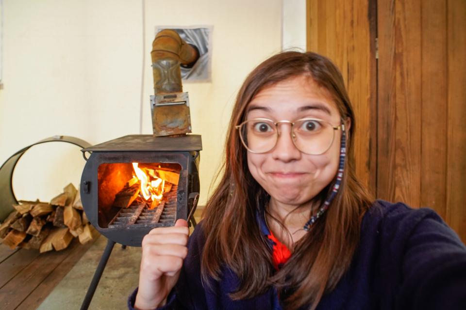 The author looks into the camera while pointing at a fire inside a small open oven behind her