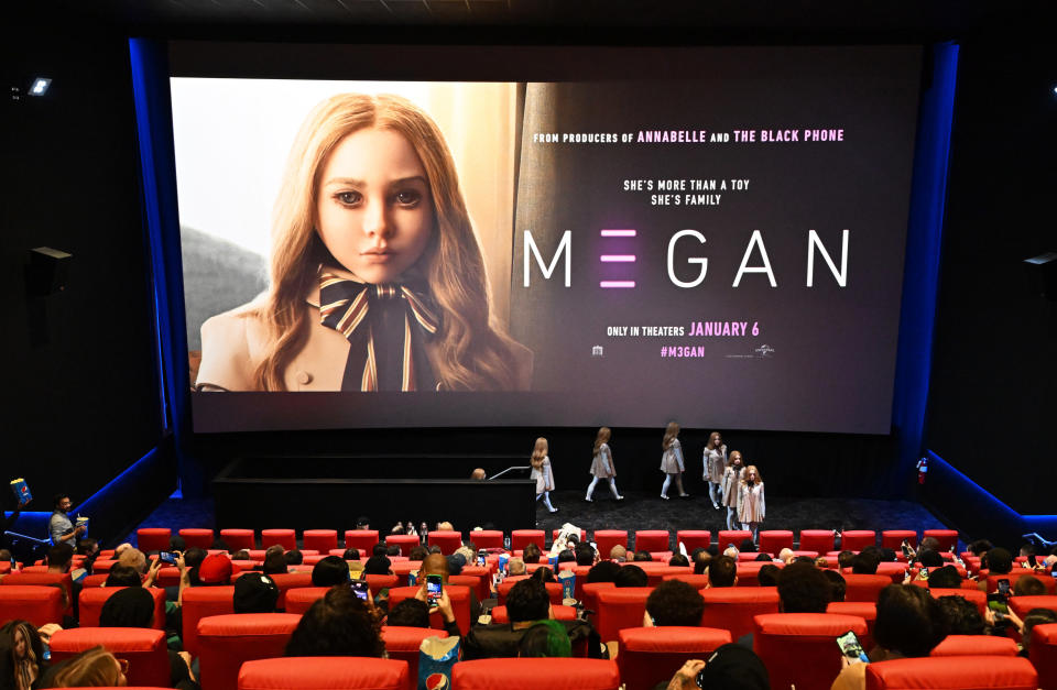 A photo of a press junket for M3GAN, with a large poster of the film showing the doll's eerie face on stage