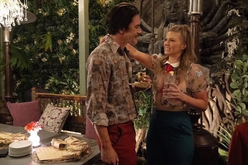 A man and woman stare and smile at one another next to a table with foliage in the background