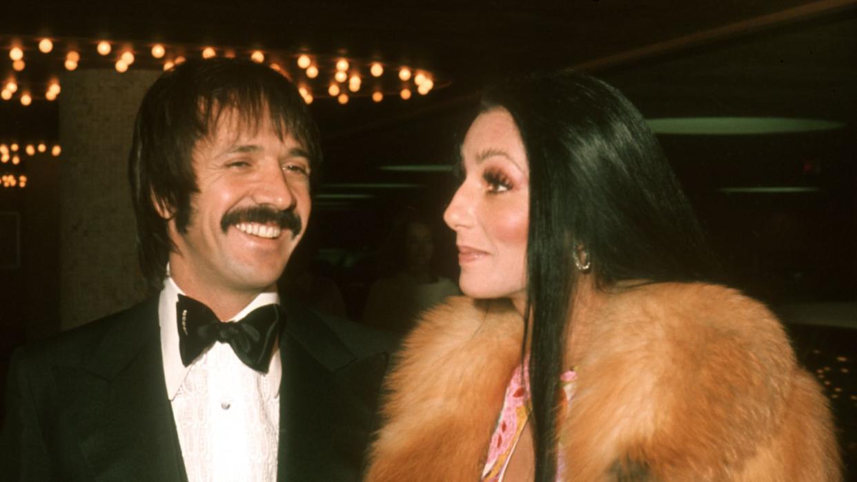 sonny and cher stand next to each other in formal attire, he smiles and she looks left with a small smile