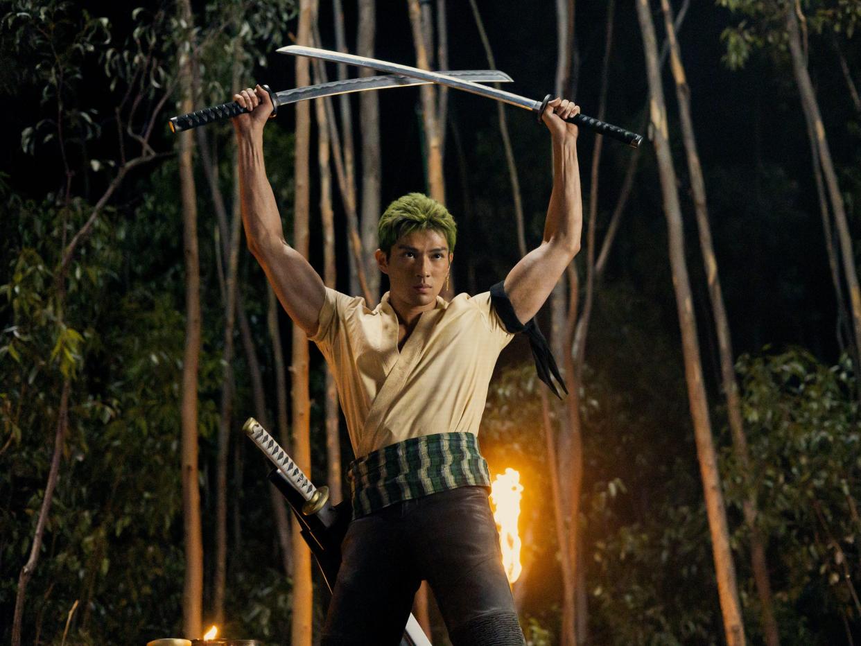 mackenyu as zoro in one piece. he's a man with short green hair, a haramaki sash around his waist, and a black cloth tied around his bicep. he's holding two swords in the air, with one more sheathed at his hip