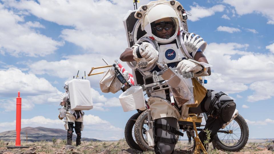  Two astronauts in the field in simulated spacesuits. the one in front uses a lunar rover and tools to pick up a soil sample. clousd and blue sky are visible in the background. 