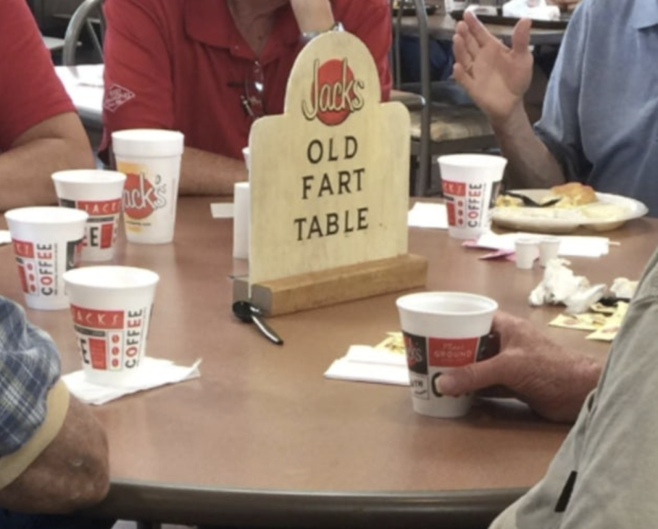 A group of seniors socializing at a table with a humorous "Jack's Old Fart Table" sign