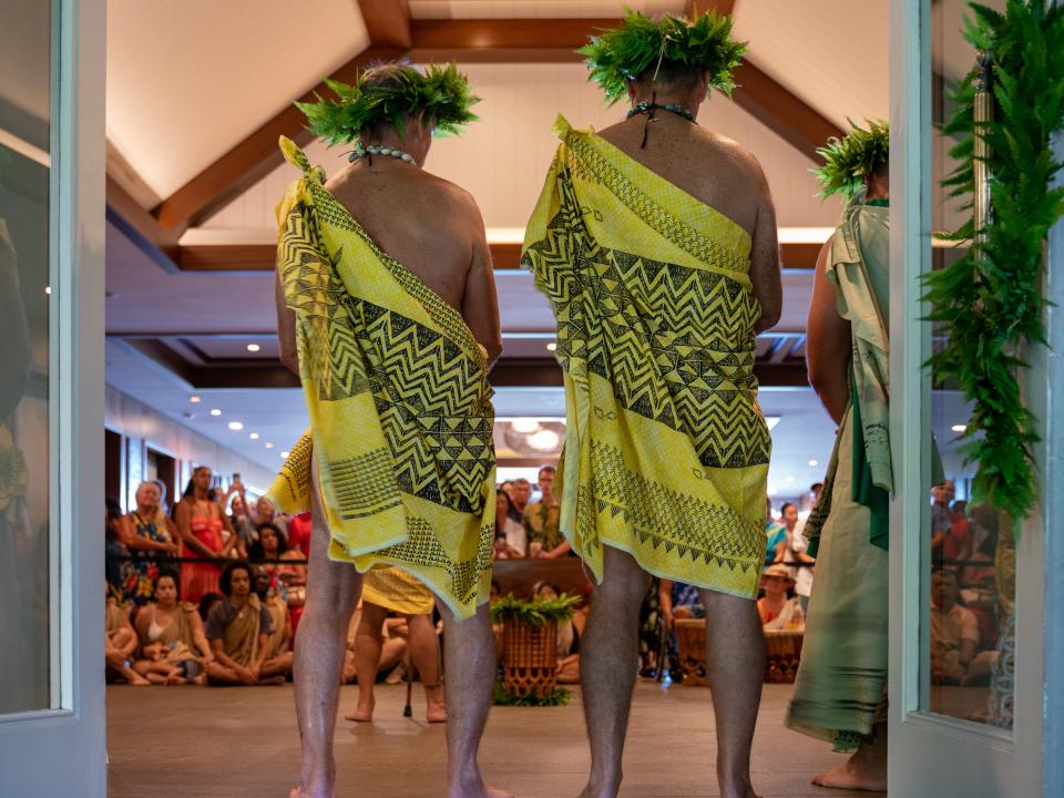 Men in traditional Hawaiian attire address a group of travelers.