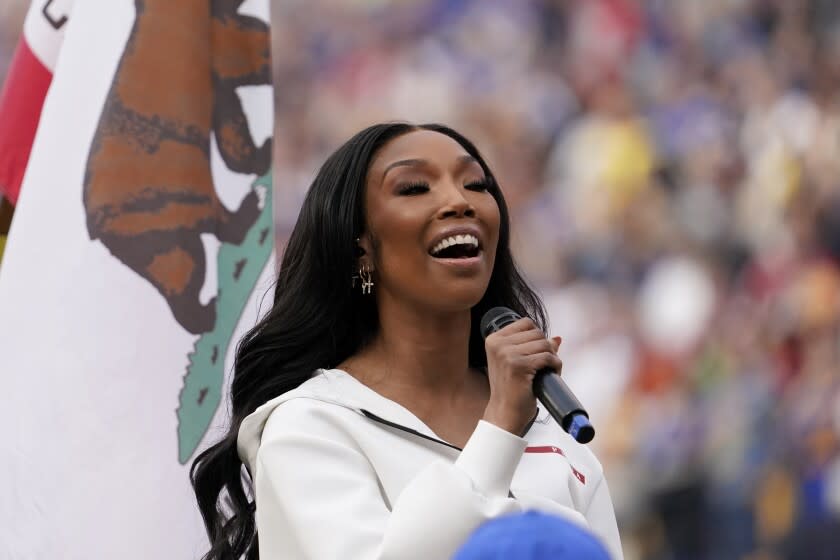 A woman with dark hair sings while holding a microphone in front of the California flag