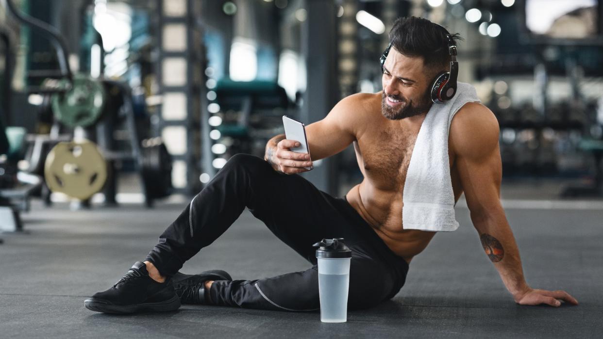  Man sitting on the floor of a. gym post-workout holding his phone smiling 