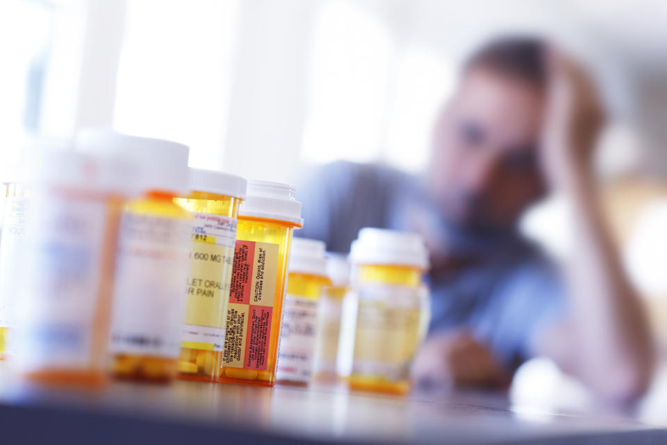 A large group of prescription medication bottles sit on a table in front of a distraught man who is leaning on his hand as he sits at his dining room table.  The image is photographed with a very shallow depth of field with the focus being on the pill bottles in the foreground.