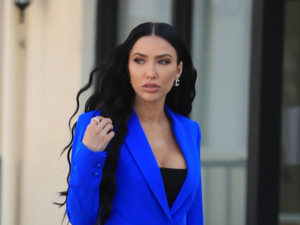 Bre Tiesi wearing a bright blue pantsuit and a black undershirt with long and wavy hair and she has her hand near her shoulder as she looks to the side