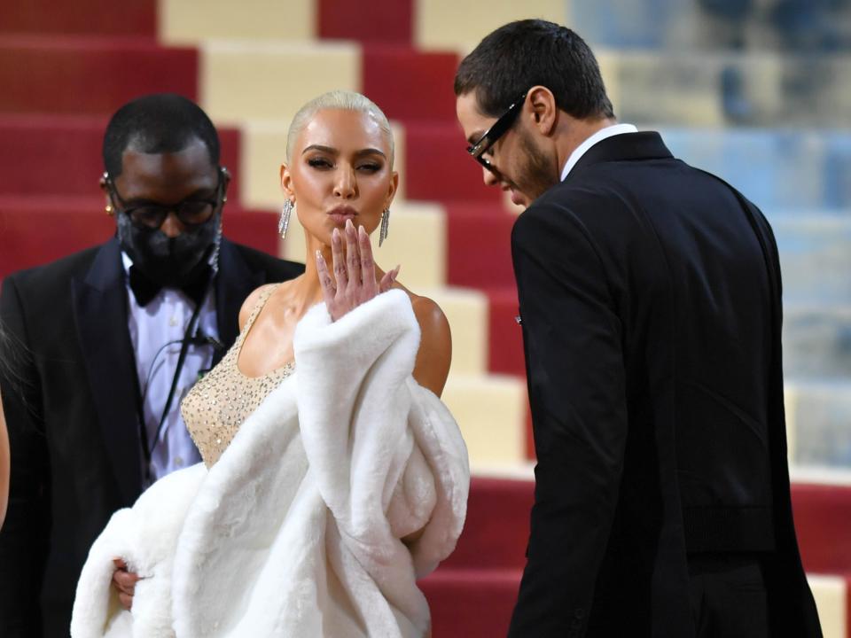kim kardashian wearing a sparkling nude dress and fur coat, blowing a kiss towards the camera, while pete davidson looks over her shoulder