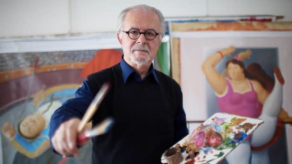 fernando botero holding a palette and two brushes while standing in front of large colorful paintings, he wears a dark sweater and collared shirt underneath with his signature black round glasses
