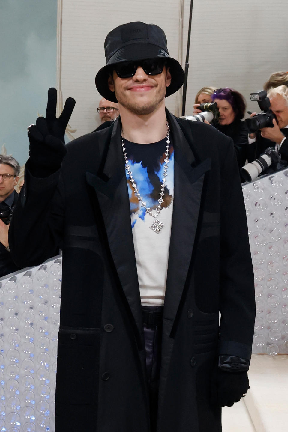 Pete giving the peace sign as he stands at a red carpet event. He's wearing a long coat, gloves, bucket hat, sunglasses, and a chain