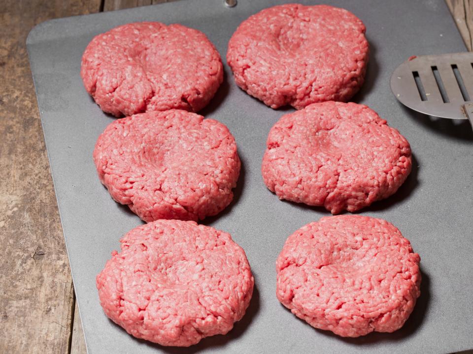 Six raw hamburgers with a thumb imprint in the middle and a spatula next to them