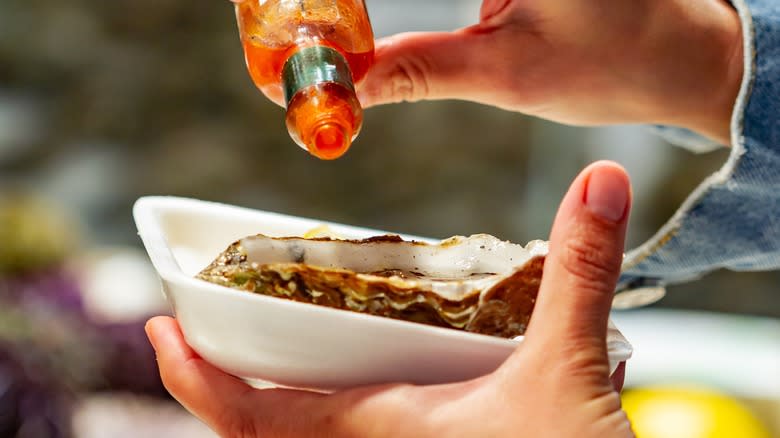 Person pouring hot sauce on oyster in shell on a dish