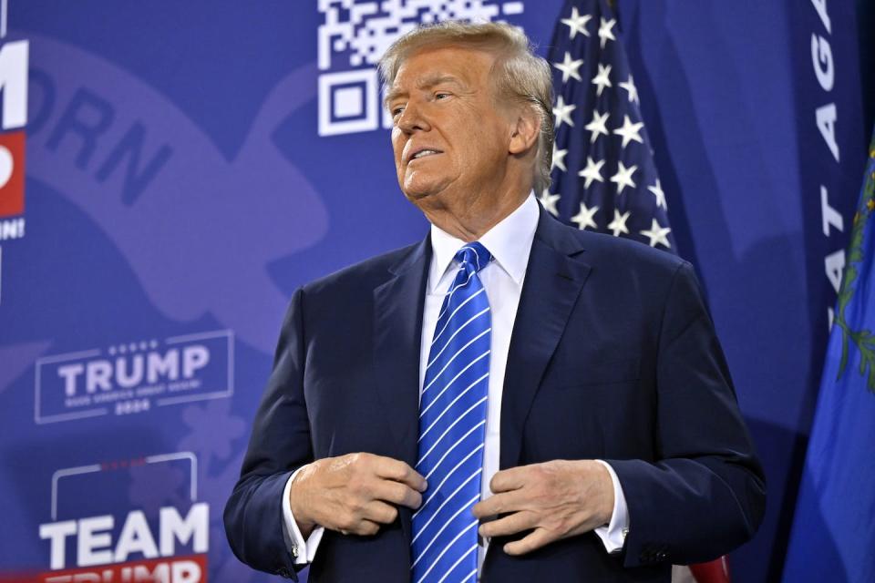 A man in a blue blazer, blue striped tie and white shirt in front of an American flag.