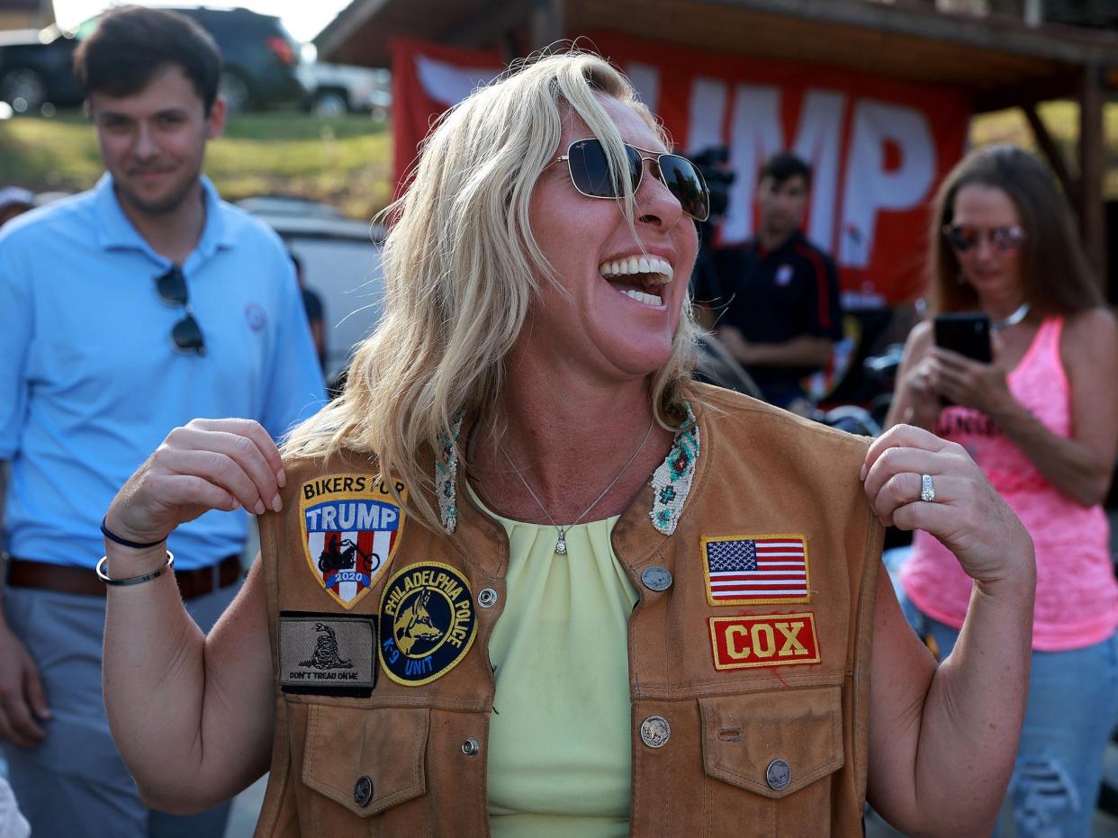 Marjorie Taylor Greene wearing a biker vest in front of a big Trump sign.