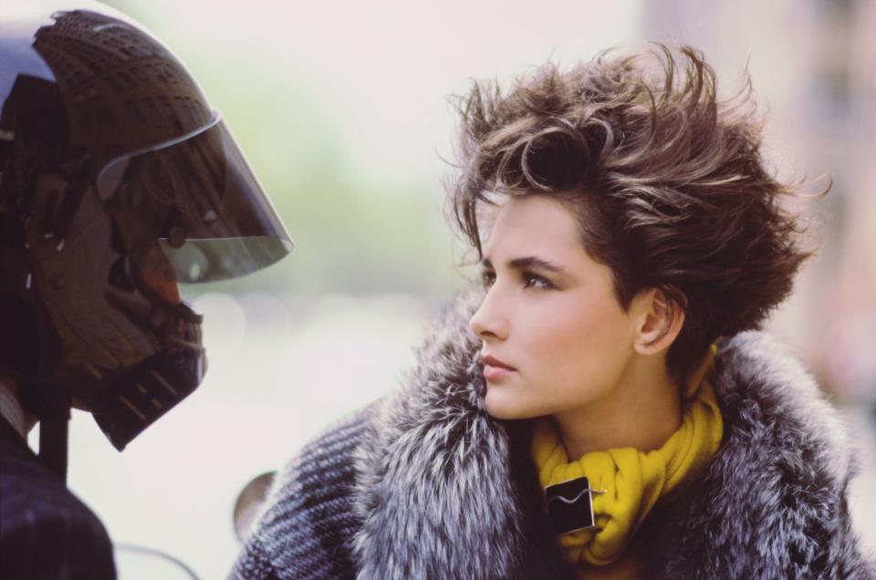 Headshot of model Daniela Ghione looking at another figure wearing on a black motorcycle helmet. Her hair is cut short in soft layers built up with product to give volume and fullness by Louis Alonzo for Nubest & Co. Makeup by Margaret Avery using Christian Dior products including Ingenue Lipstick and Light Play Eyeshadows. Ghione is wearing a black and grey plaid wool and mohair jacket with a silver fox collar over a cashmere and angora yellow sweater by Emanuel Ungaro. Accessories: pin and necklace by Laurence de Vries. Man's outfit by Giorgio Armani and Helmet by Simpson.