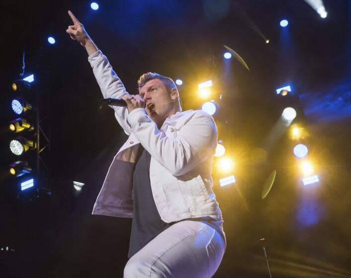 Nick Carter is wearing a white jacket and pants while performing on stage with microphone to mouth, finger pointing up