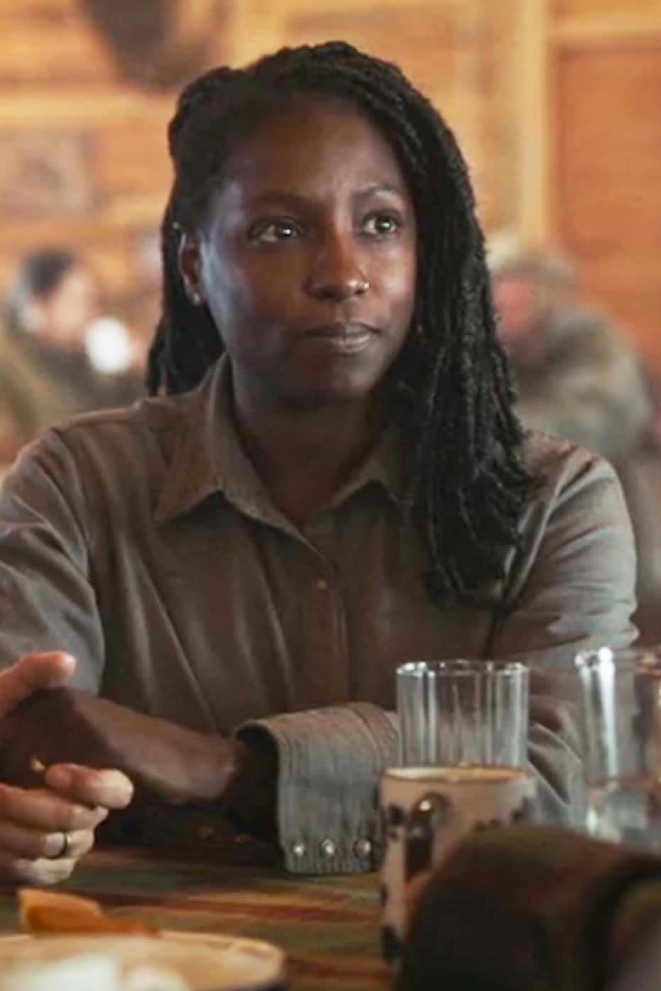 Maria in The Last of Us Season 1 sitting at a table, appearing engaged in conversation, with dreadlocked hair and a buttoned shirt