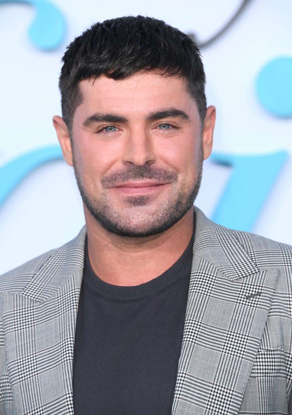 Zac Efron smiles for the camera at an event, wearing a checkered blazer over a dark shirt