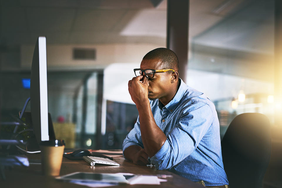 Man in glasses at a desk appears stressed or concentrating with a hand on his forehead near a computer