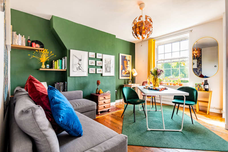 Dining room with turquoise table with white legs, green and rose velvet dining chairs, green area rug, one deep green accent wall, window with gold curtain, small gold shelf with liquor, wine, oval gold frame mirror, framed art. To the left of the dining area is a large gray sofa with blue and maroon velvet throw pillows