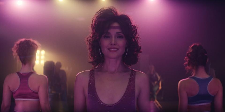 Rose Byrne in a leotard, narrow headband, large hoop earrings and a perm.