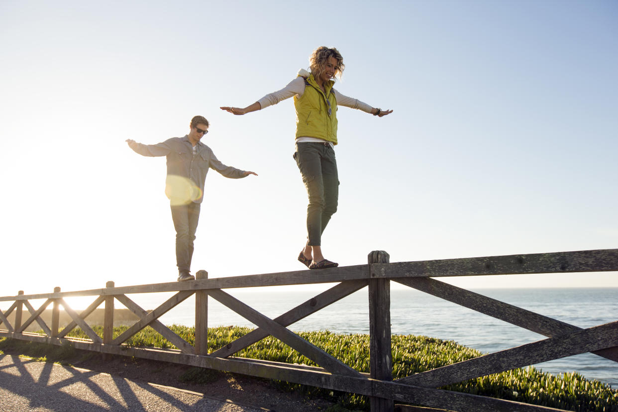 A man and a woman walking along the top of a fence with their arms extended.