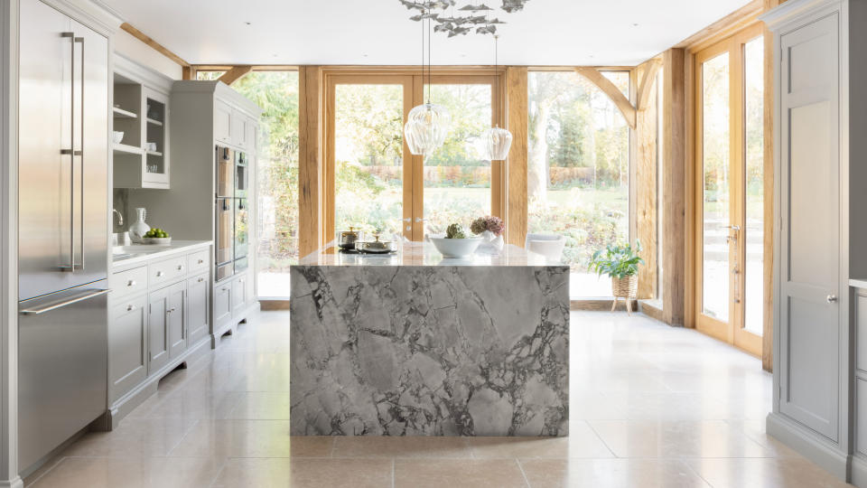 Large white kitchen space with statement marble island, wooden joinery and door frames, large stone floor tiles, white kitchen cabinets, decorative hanging feature and lights over island