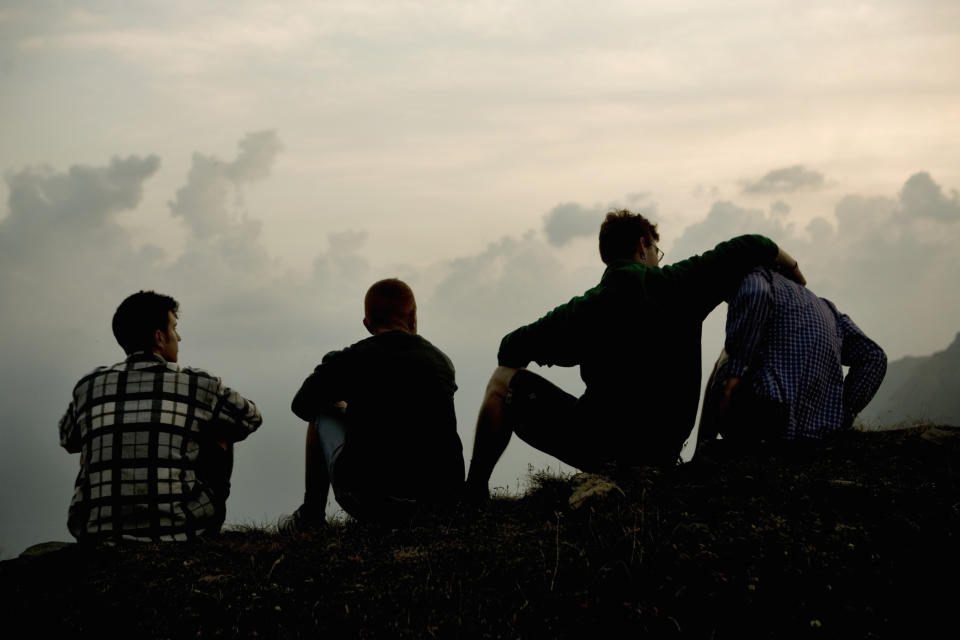 Four people sit on a hilltop, gazing into the distance with their backs to the camera, appearing relaxed and contemplative