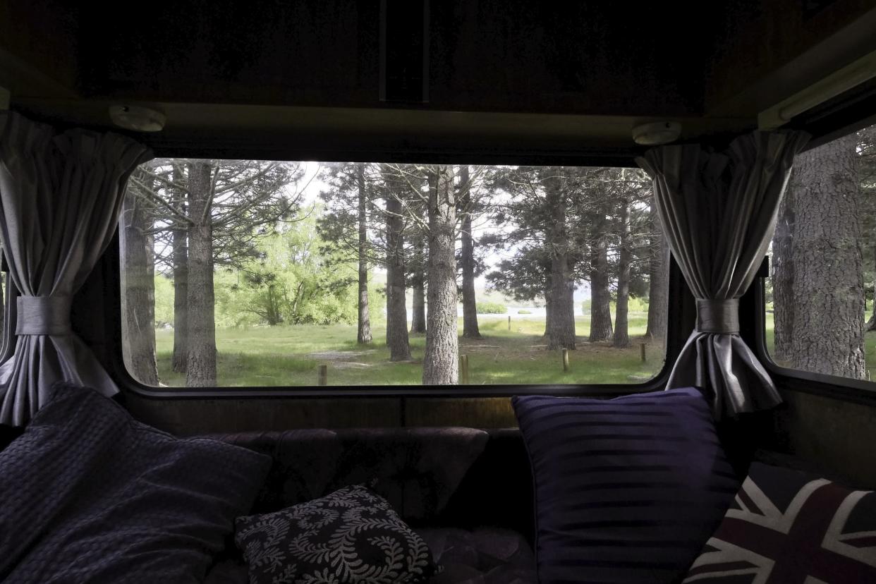 Forest view from back window of vintage motor home, throw cushions on the couch and the curtains open.
