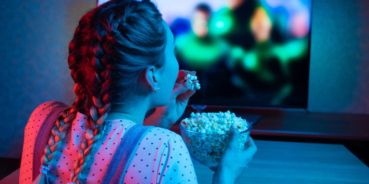 A young girl watching movies and eating popcorn with a bowl on the background of the TV. The color bright lighting, blue and red. Relax, rest at home when watching TV, film. Background for design