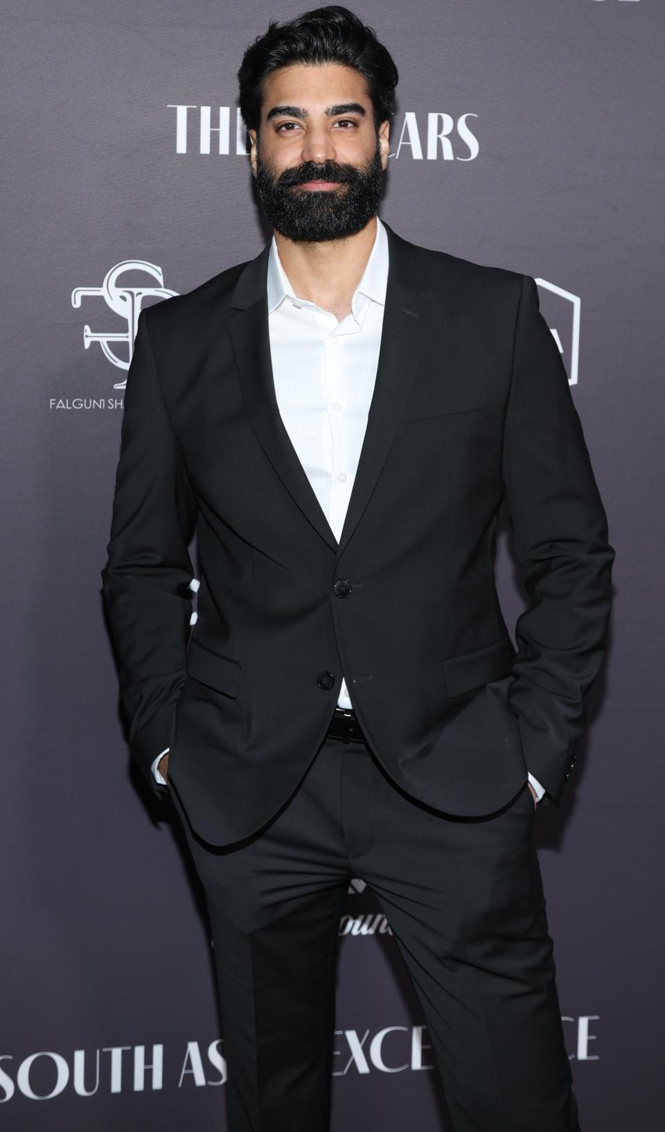 Raymond Ablack in a tailored suit standing against a branded backdrop at an event